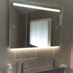 Badkamerspiegel van 100cm breed en 70cm hoog, in een grijs betegelde badkamer met en donkergrijze wastafel en zilveren kranen.